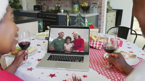 pareja afroamericana con vino usando computadora portátil para la videollamada de navidad con familia feliz en la pantalla