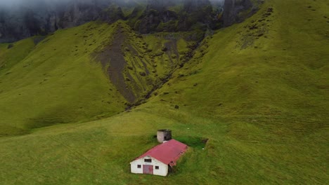 single lonely house at base of the mountain in icelands countryside with low clouds and rolling meadows around, remote summer cottage