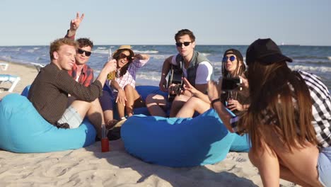 Young-hipster-girl-recording-a-video-or-taking-pictures-of-group-of-friends-sitting-on-easychairs-on-the-beach,-playing-guitar-and-singing-on-a-summer-evening.-Slowmotion-shot