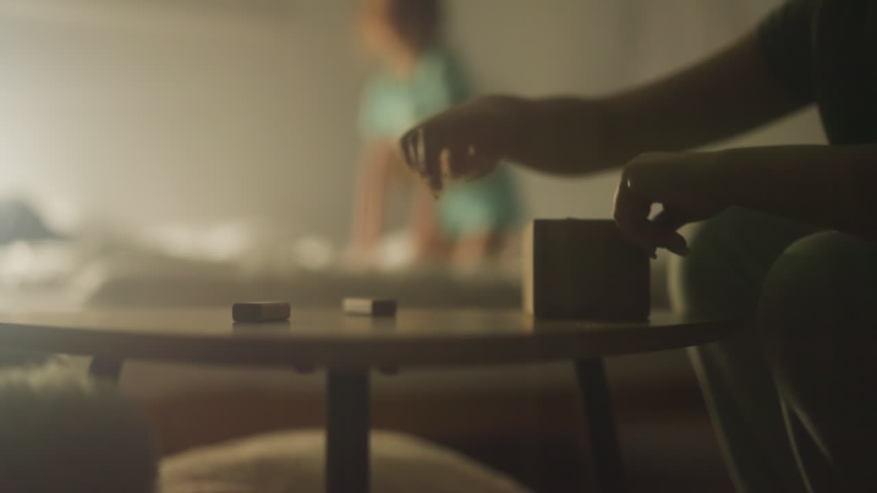 딸이 침대에서 재미를 느끼는 동안 여성은 건축 세트의 나무 부분을 카드보드 상자에 넣습니다. 엄마는 잠자리에 들기 전에 테이블에서 남은 게임을 청소합니다.