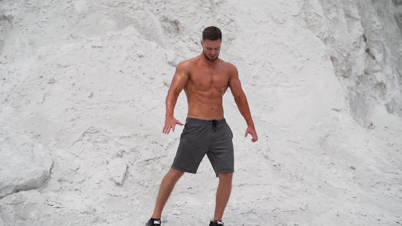 Athletic man with trained body outdoors. Muscular sportsman shirtless in shorts posing to camera on white rocky background.
