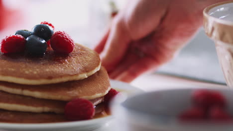 Woman-Putting-Stack-Of-Freshly-Made-Pancakes-With-Maple-Syrup-And-Berries-On-Table-For-Pancake-Day