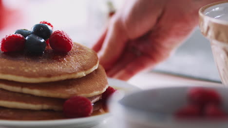 woman putting stack of freshly made pancakes with maple syrup and berries on table for pancake day