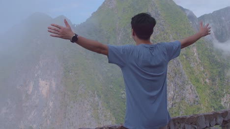 asian hiker male standing on a veranda cliff and raising his hands celebrating reaching up top of foggy mountain