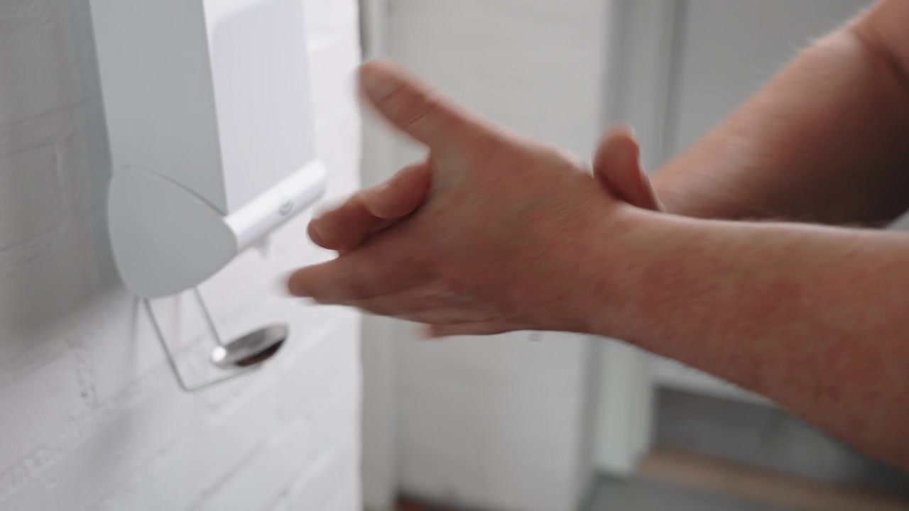 Primer plano de una persona aplicando desinfectante de manos desde un dispensador montado en la pared, mostrando la higiene diaria de manos, desinfección y prevención de infecciones en un entorno público o laboral