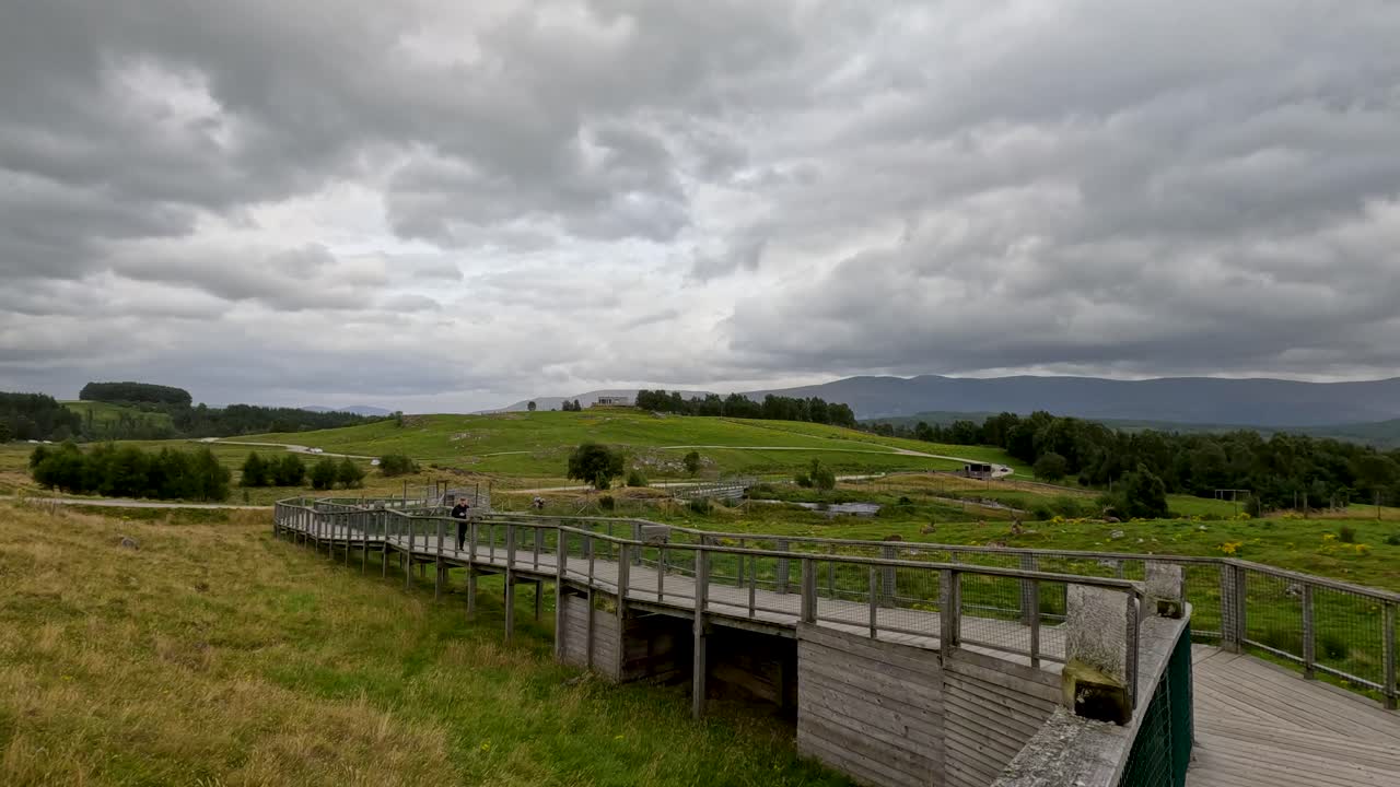 Slow pan across wooden boardwalk, grassy meadow, distant hills, under dramatic overcast sky