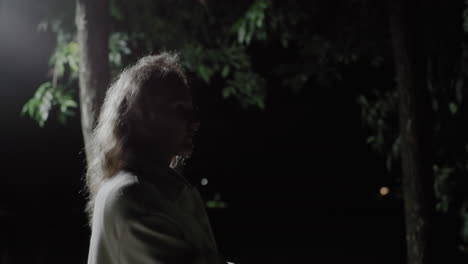 side view of woman in white coat with curly hair walking away from bright street light at night, silhouette fading into darkness among tall trees