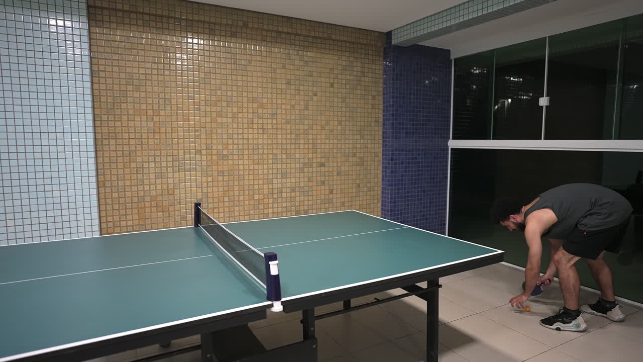 A man playing table tennis indoors, captured mid-action as he returns the ball. The scene features a green ping pong table with a net and a tiled wall background, a casual and sporty atmosphere