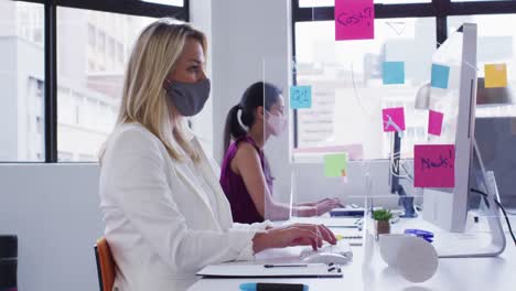 Diverse-businesswomen-wearing-face-masks-working-at-desks-in-office-with-hygiene-screen-between-them