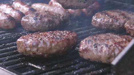 chef flipping juicy sizzling burgers on hot barbecue with sausages in background. summer outdoor high protein slow motion food. smoke rising from red hot charcoals beneath iron grill.