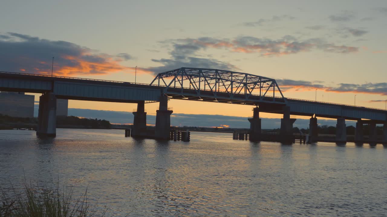 A striking sunset casts warm light over a steel truss bridge in New Jersey, reflecting across the river in a tranquil evening scene.