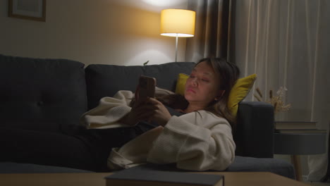 woman spending evening at home lying on sofa with mobile phone scrolling through internet or social media 1