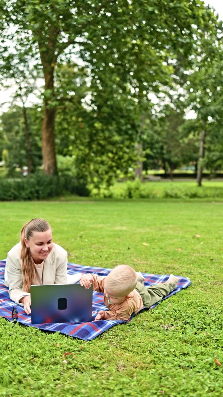 En ung mor sidder på græsset i en park og arbejder på en bærbar computer, mens hendes barn løber rundt. En mor arbejder hjemmefra under barselsorlov i naturen, mens hendes barn leger i nærheden.