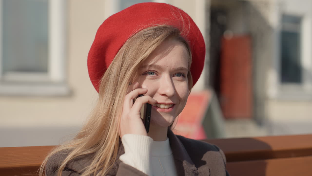 Giovane donna caucasica su una panchina che parla al telefono, con un basco rosso e i capelli al vento, espressione pensierosa, sotto una luce dorata e soffusa, con architettura urbana e pedoni in lontananza, in un'atmosfera contemplativa e artistica.