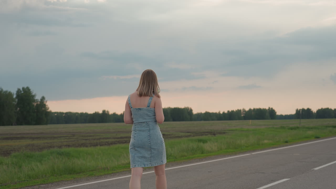 Rear view of woman waving colourful pinwheel while walking along straight asphalt road beside open grassy field under cloudy sky hair swaying in breeze