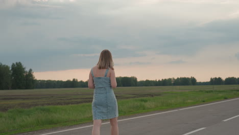 rear view of woman waving colourful pinwheel while walking along straight asphalt road beside open grassy field under cloudy sky hair swaying in breeze