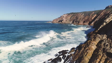 Aéreo---Volando-Desde-La-Costa-Rocosa-Sobre-Fuertes-Olas-Que-Se-Estrellan-Contra-Acantilados-Escarpados,-El-Punto-De-La-Bahía-De-Mossel