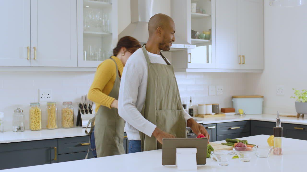Coppia multirazziale che cucina insieme in cucina, sorridendo e usando un tablet per la ricetta, a casa.