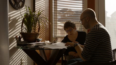a senior woman sits at a kitchen table with a middle-aged man, in a warmly sunlit house . he helps her fill out a paper questionnaire. she answers, as he writes with a pen. both wear reading glasses.
