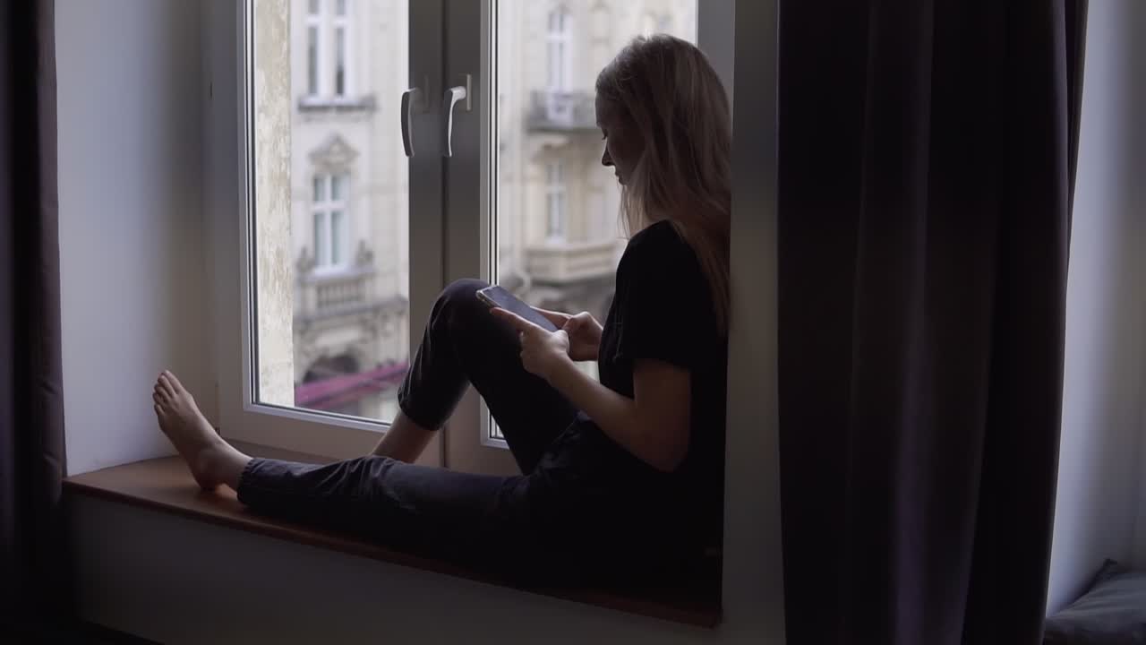 Rare view of a woman sitting on window sill and using smartphone at home