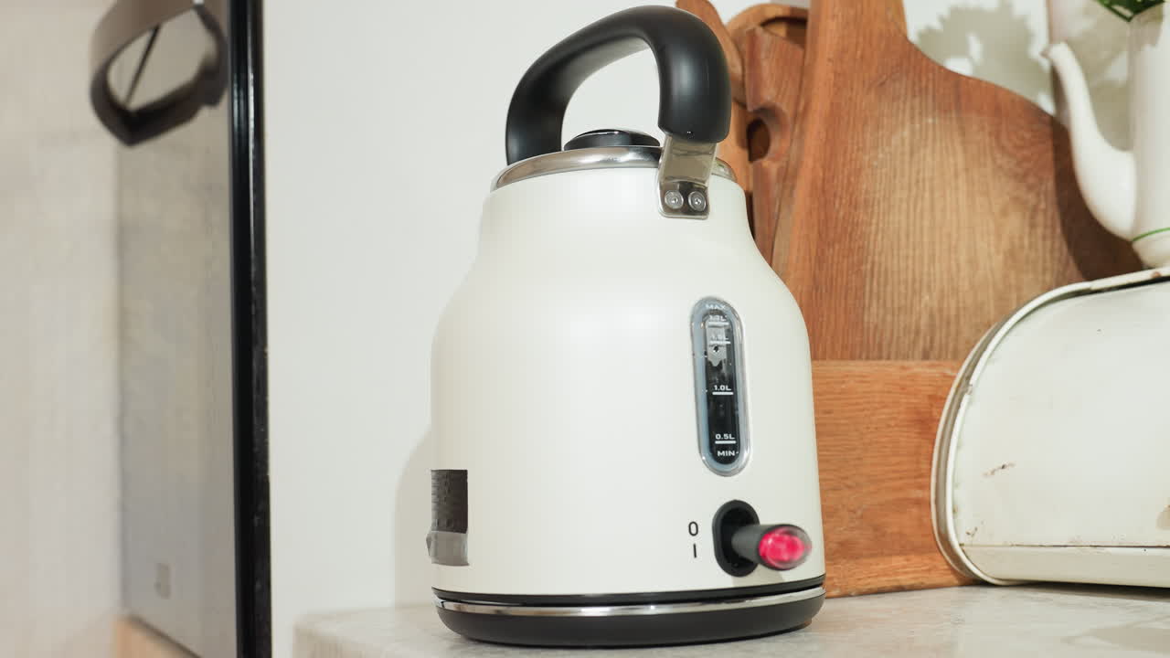 White electric kettle with water steaming inside and blinking indicator light sits on kitchen countertop beside wooden spatula and vintage bread box while slightly shaking during boiling process