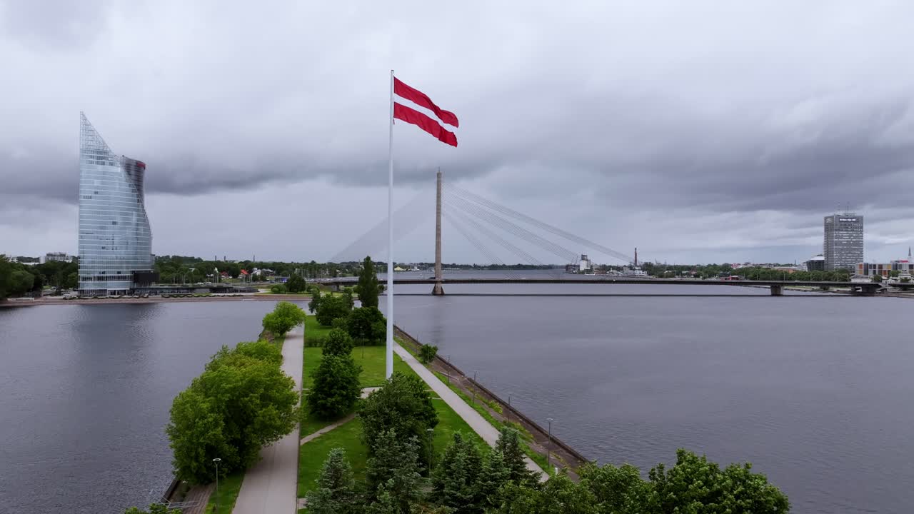 Majestætisk rødt flag vajende i vinden, mens drone svæver over Rigas flodpromenade.
