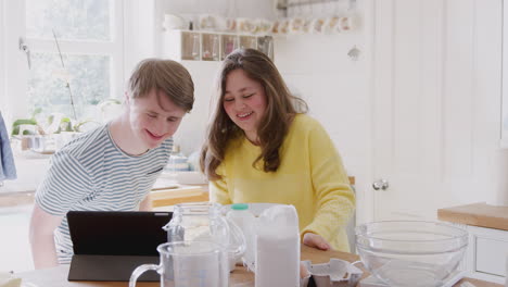 Una-Pareja-Joven-Con-Síndrome-De-Down-Sigue-Una-Receta-En-Una-Tableta-Digital-Para-Hornear-Pasteles-En-La-Cocina-De-Casa