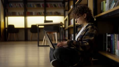 A-girl-with-curly-hair-wearing-glasses-sits-on-the-floor-near-shelves-with-books-and-takes-notes-on-a-laptop-in-the-library