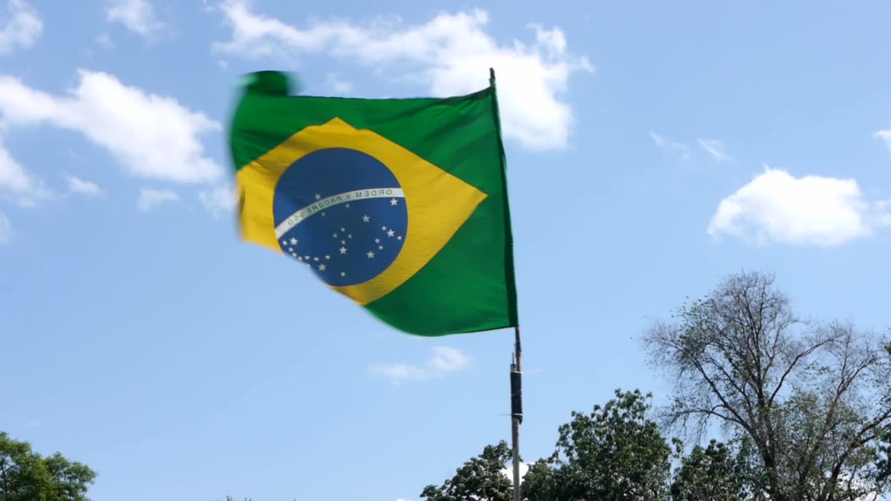 Uma bandeira brasileira tremula ao vento e está presa a um mastro improvisado. O céu está azul, nuvens podem ser vistas e as árvores se movem ao vento.