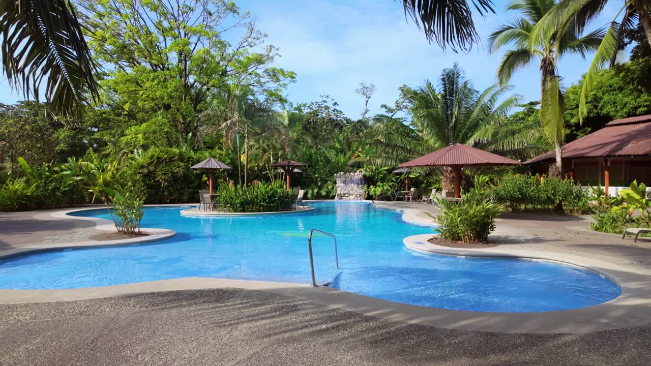 Idyllic exotic pool with a mini waterfall and an island in the middle of it surrounded by tropical plants and palm trees on a sunny day in Costa Rica.