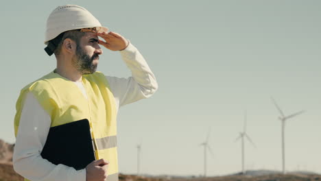 a young caucasian engineer in a white helmet and reflective vest checks wind turbines on a sunny day, emphasizing the importance of renewable energy in saving our planet