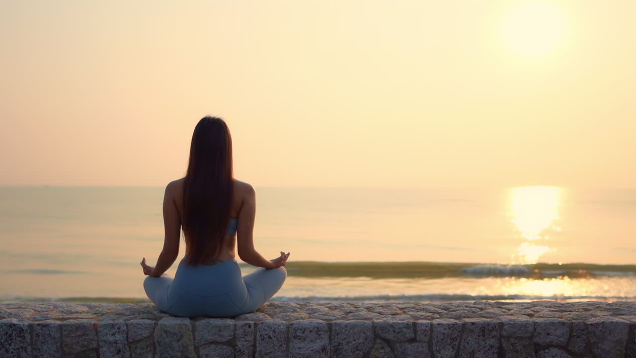 Back to camera, A young woman sits meditating on a stone wall facing the incoming tide at sunset. copy space