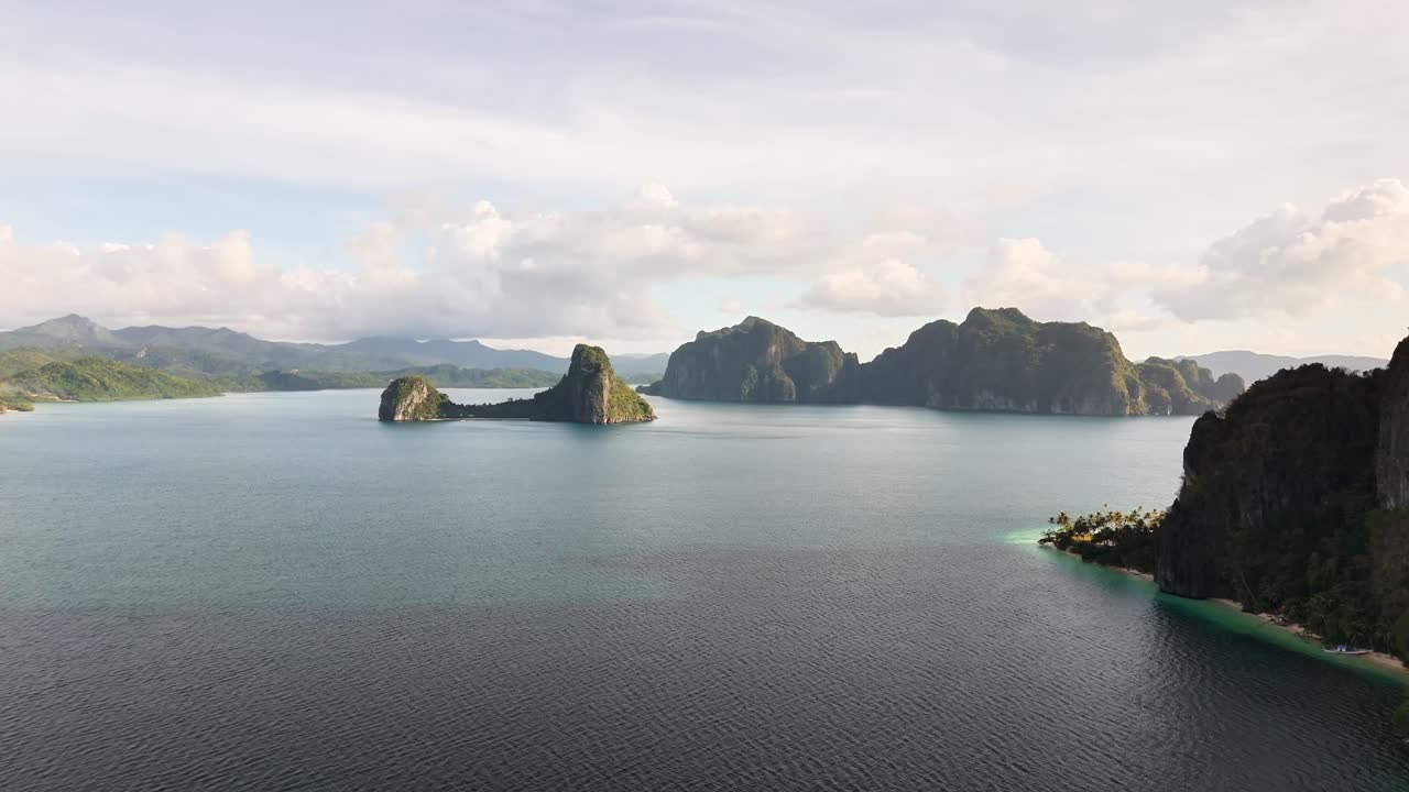 Aerial view of calm blue waters surrounding Pinagbuyutan Island in Palawan, Philippines, with dramatic limestone cliffs, lush green terrain, distant mountain ranges, and scattered tropical islets