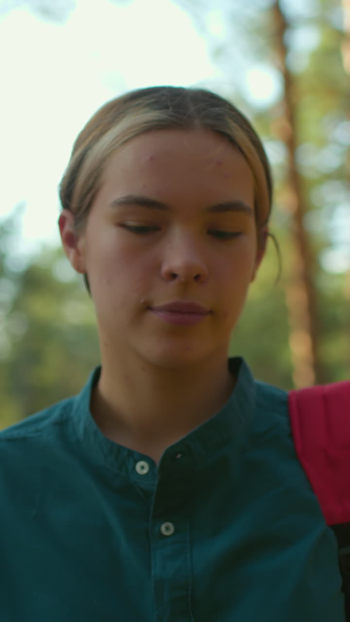 una mujer con el cabello atado hacia atrás, con camisa verde y mochila roja, camina a través del bosque iluminado por el sol con una cálida sonrisa, mira pensativo a su alrededor mientras la luz del sol se refleja suavemente en su cara