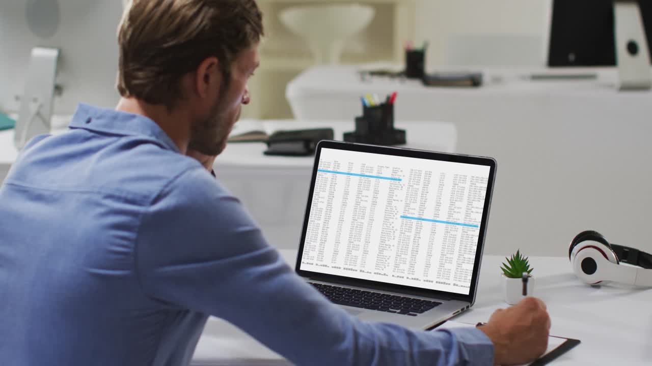Caucasian man sitting at desk processing on laptop and making notes