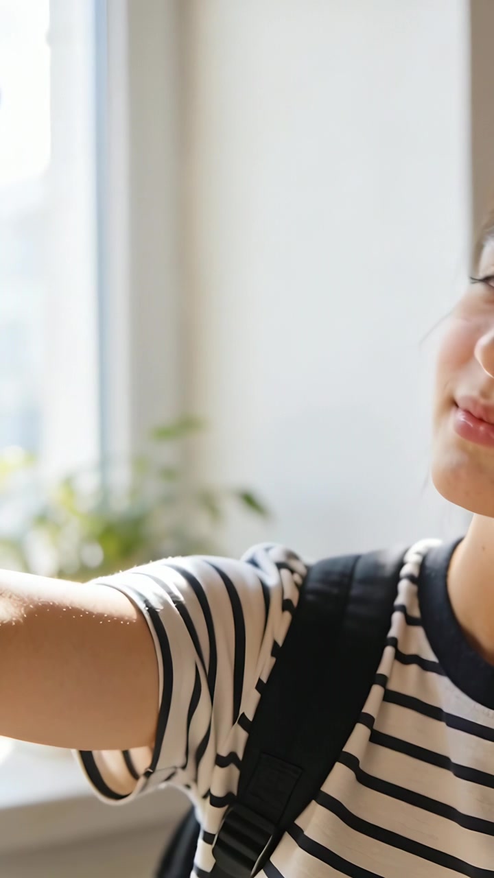 Vertical video: Raising phone, teen wearing striped tee and backpack posing for selfie by window
