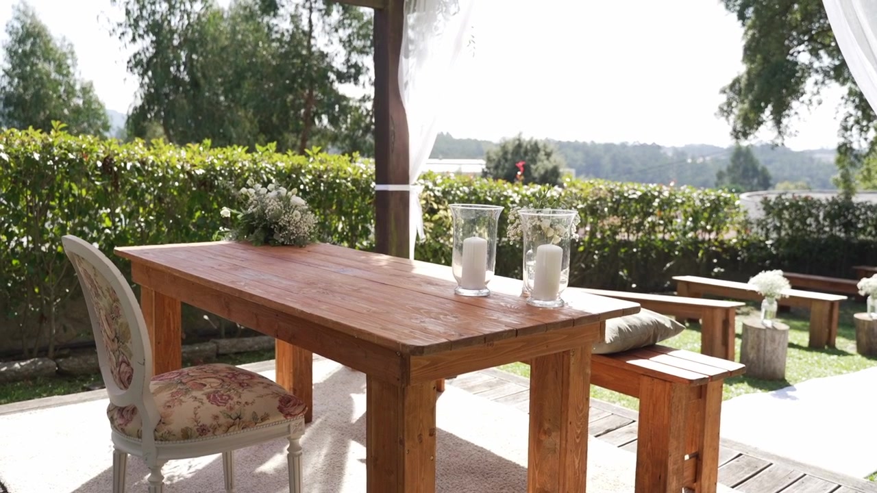 Wooden table with candles and chairs arranged outdoors near garden and water
