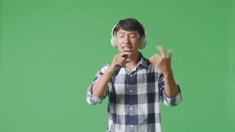 young asian teen boy wearing headphone and holding a microphone and singing on the the green screen background