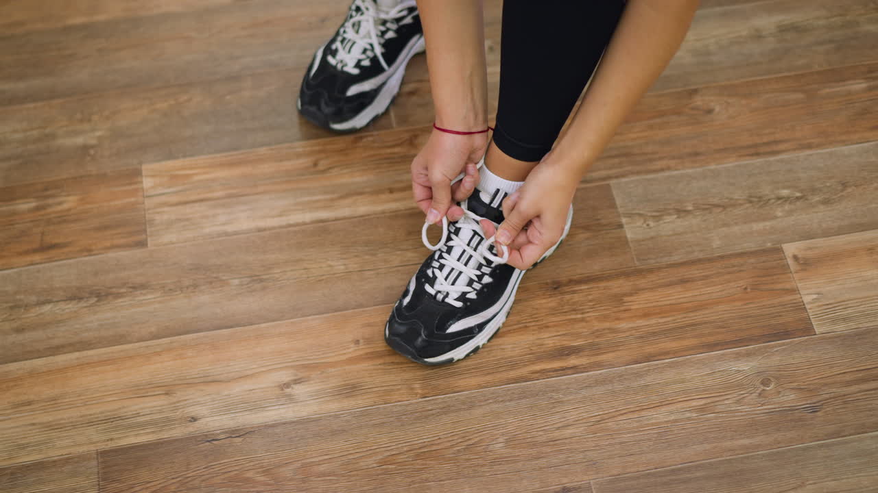 Signora con leggings neri che si china per allacciare le scarpe da ginnastica bianche e nere su un pavimento di legno, concentrandosi sulle scarpe e sul movimento della mano, mostrando attenzione e cura nei dettagli