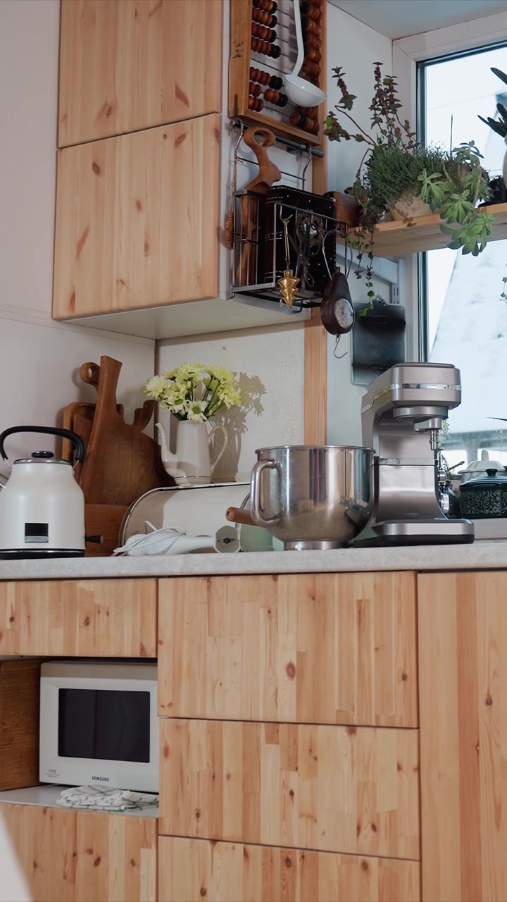 Close up of kitchen countertop with white kettle metal mixing bowl. White vase with yellow flowers and wooden cutting boards are. Natural light from window illuminates scene