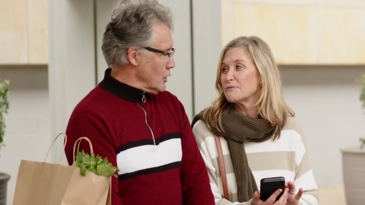 Senior couple walking along facade carrying grocery bag, glancing, talking and checking smartphone