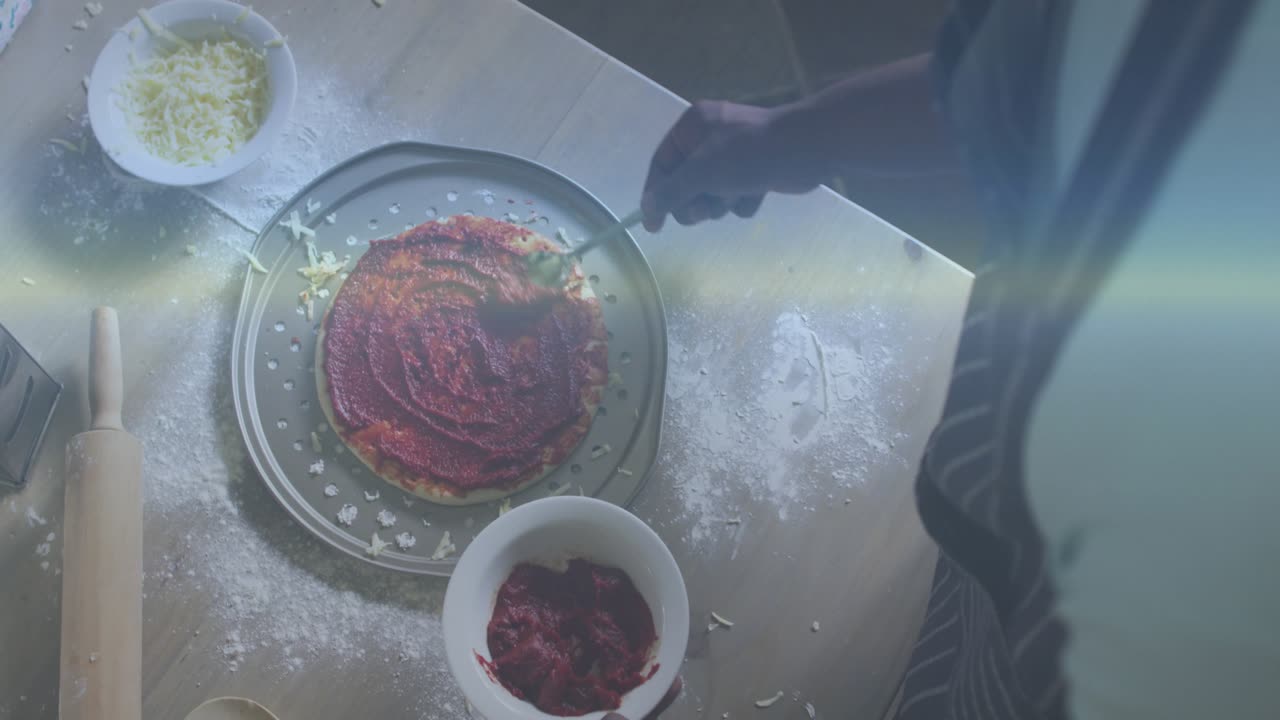 Man scooping tomato sauce and spreading concentric rings on pizza crust in pan for cooking