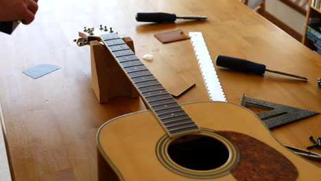 Close up hands of a luthier sanding and leveling the frets on an acoustic guitar neck fretboard on a wood workbench with lutherie tools
