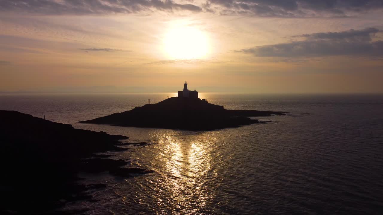 Aérea descendente do Farol de Mumbles ao nascer do sol com sombra projetada no mar. Reflexo dourado do sol no oceano com nuvens dinâmicas. Perfeito para turismo, documentários de viagem. País de Gales, Reino Unido. 4K.