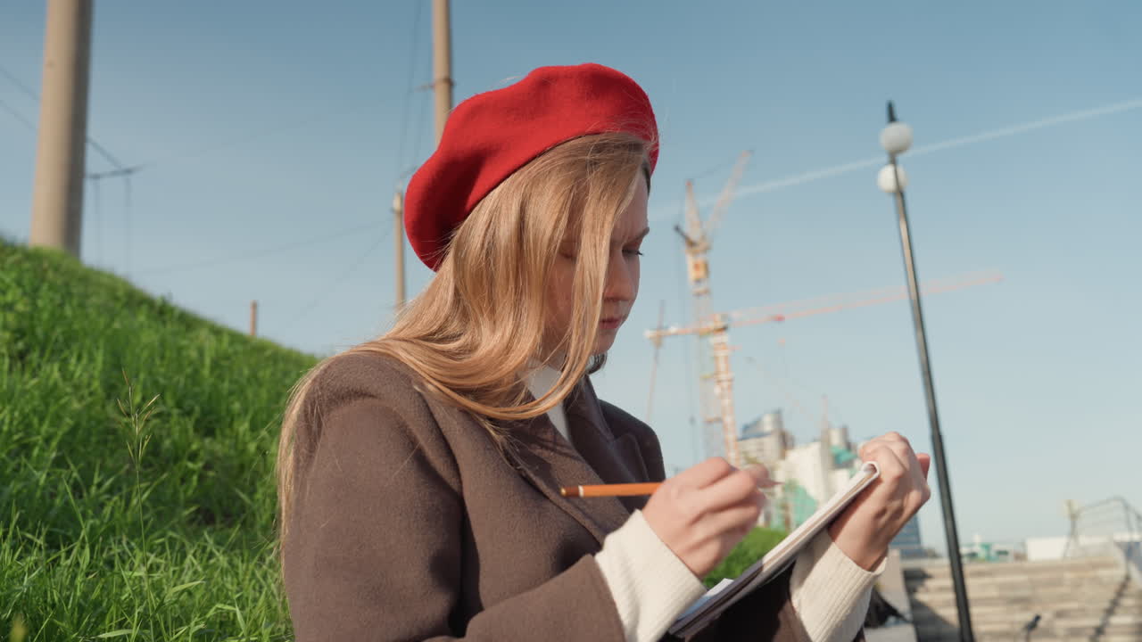 White woman sketching city with pencil, red beret and wool coat, focused hands drawing lines in notebook, green slope and construction cranes nearby, sidewalk and lamppost, creative outdoor session