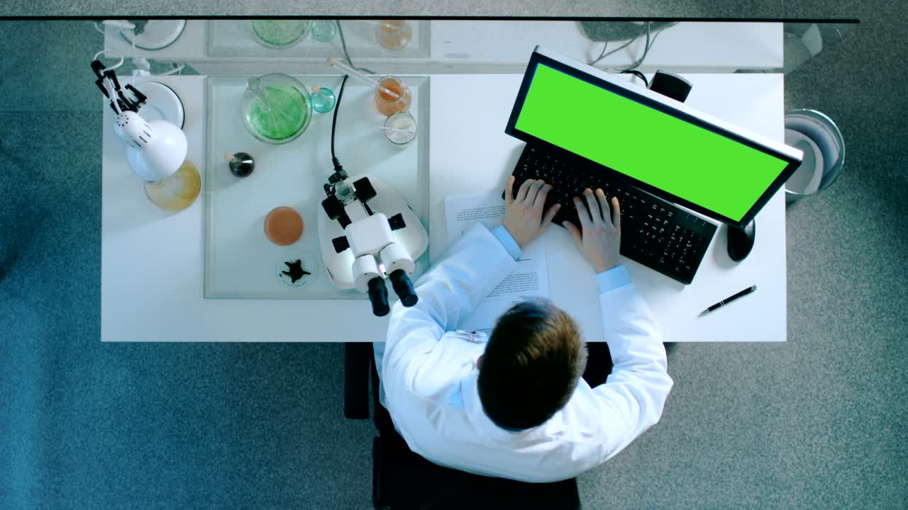 Top View of a Scientist Working on a Personal Computer with Green Screen on, Looking Into Microscope and Writing Down Results on a Document with Charts.