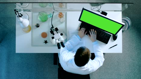 vista superior de un científico trabajando en una computadora personal con pantalla verde encendida, mirando al microscopio y escribiendo los resultados en un documento con gráficos.
