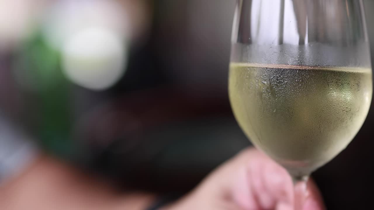 Una mano tiene delicatamente un calice da vino ghiacciato riempito di vino bianco su uno sfondo sfocato.