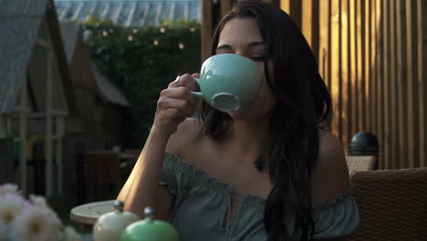 Portrait-of-a-young-hispanic-latina-good-looking-woman-drinking-coffee-or-tea-and-smiling-while-sitting-outdoor