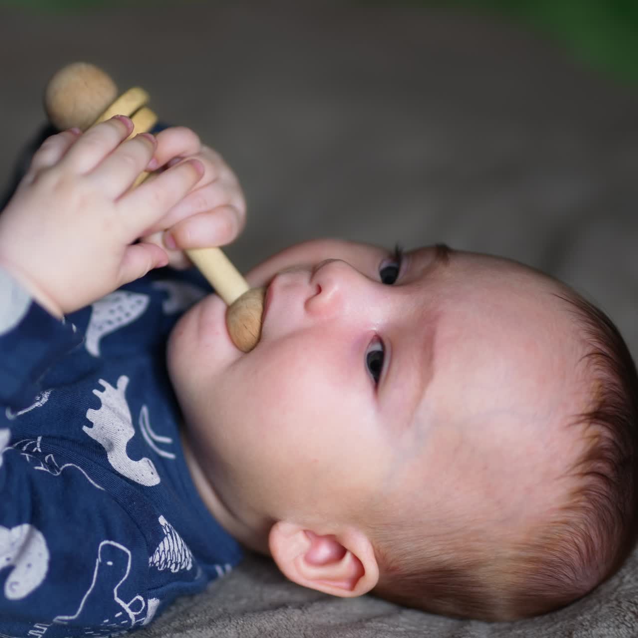 Little boy in blue clothes lies on back holding a wooden toy in hands. Tiny boy puts a toy in his mouth. Close up